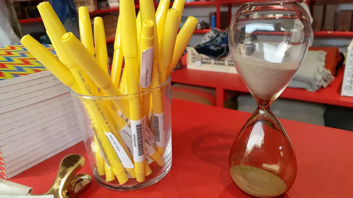 Glass hourglass with golden sand beside yellow pens on a red counter
