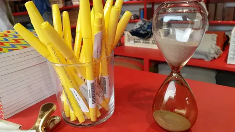 Glass hourglass with golden sand beside yellow pens on a red counter